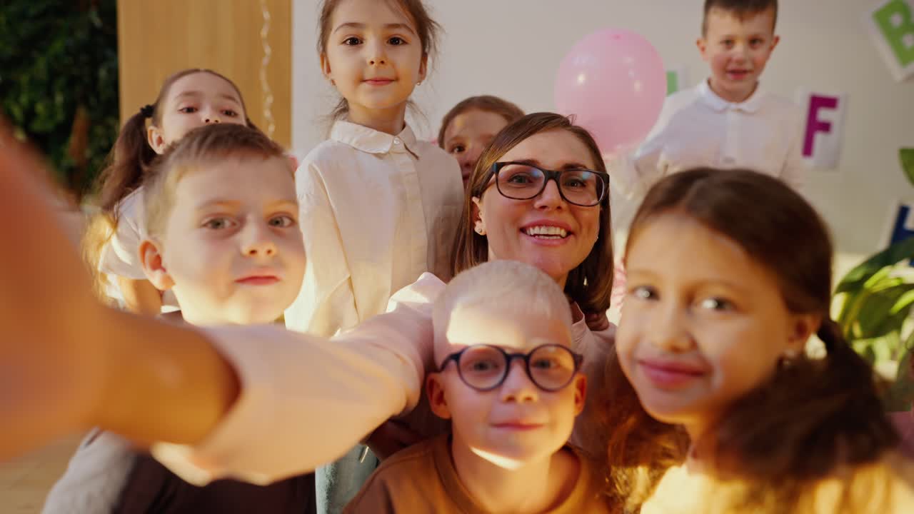 A happy teacher with a bob hairstyle in glasses and a white shirt takes a selfie with her students, preschool children in a club for preparing children for school