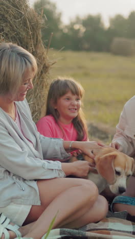 abuela e hija con niños jugando con un perro con correa al aire libre cerca de balas de heno, cálida reunión familiar en el campo durante la puesta de sol, ocio rural casual