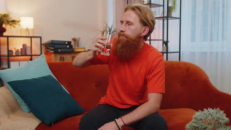 Thirsty-young-adult-man-sitting-at-home-holding-glass-of-natural-aqua-make-sips-drinking-still-water