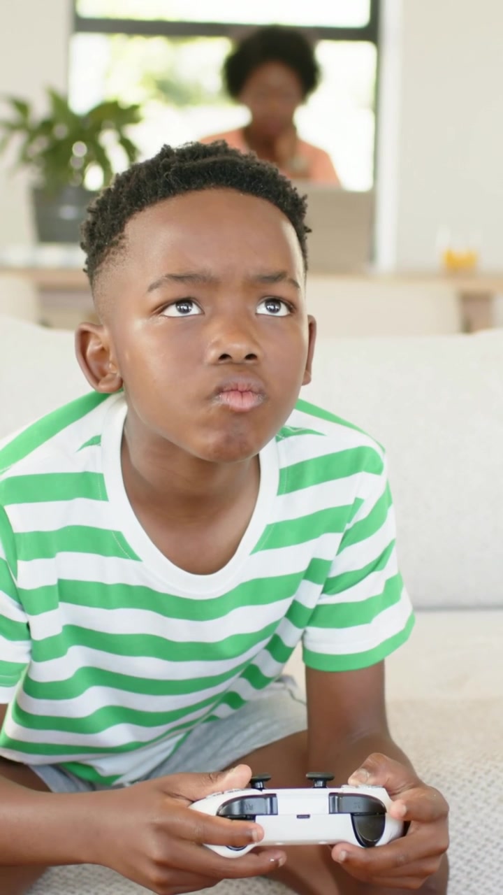 Bambino seduto sul divano con un controller bianco che preme i pulsanti giocando, mentre la mamma lavora al laptop.