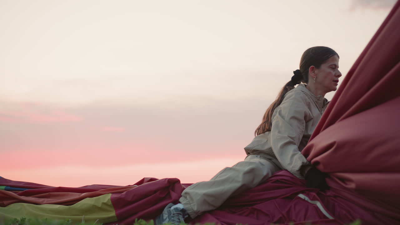 woman kneeling on deflated hot air balloon fabric on green field at sunset grasping red envelope panel under soft pastel sky during twilight countryside landing pack up operation