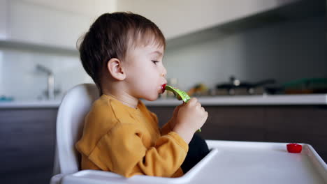 cute kid sits at high chair holding a pack with juice in his mouth. baby boy winking as he eats fruit puree. side view.