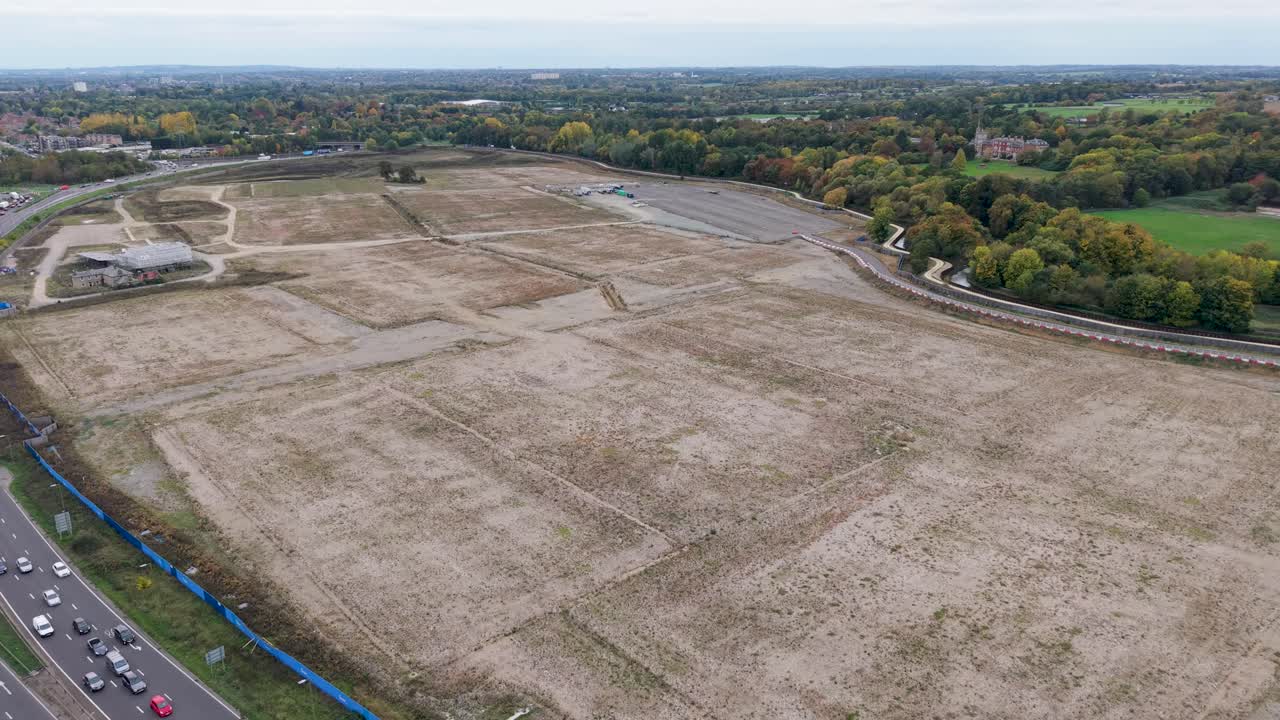 High-altitude drone view over the former site for Sunset Studios in Waltham Cross, Hertfordshire. The shot captures empty land, blue hoarding, autumn trees, and the busy A10 road junction below
