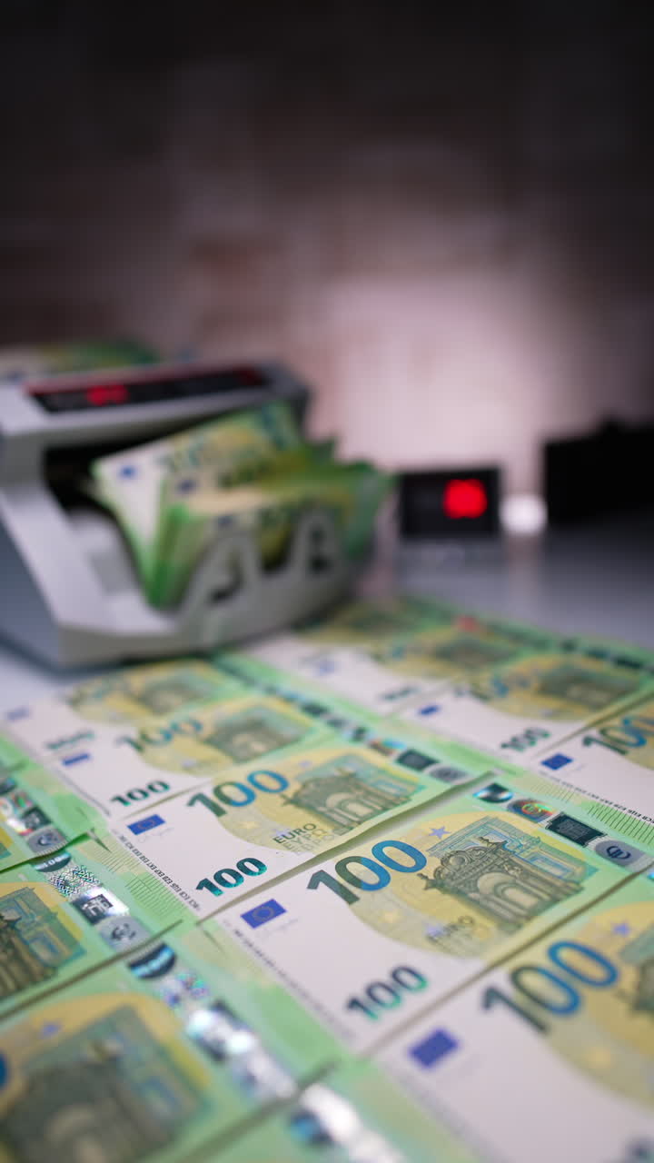 Green one hundred euro bills are laid on the desk. A stack of money is counted in the cash counting machine. Male hand puts banknotes at the foreground. Close up. Blurred backdrop. Vertical video.