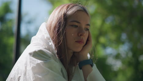 close-up of young woman in transparent raincoat, resting chin on hand in peaceful, contemplative pose, with a softly blurred green natural background