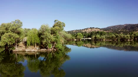 Flying-by-island-of-trees-on-mirror-lake-with-colorful-blue-reflection