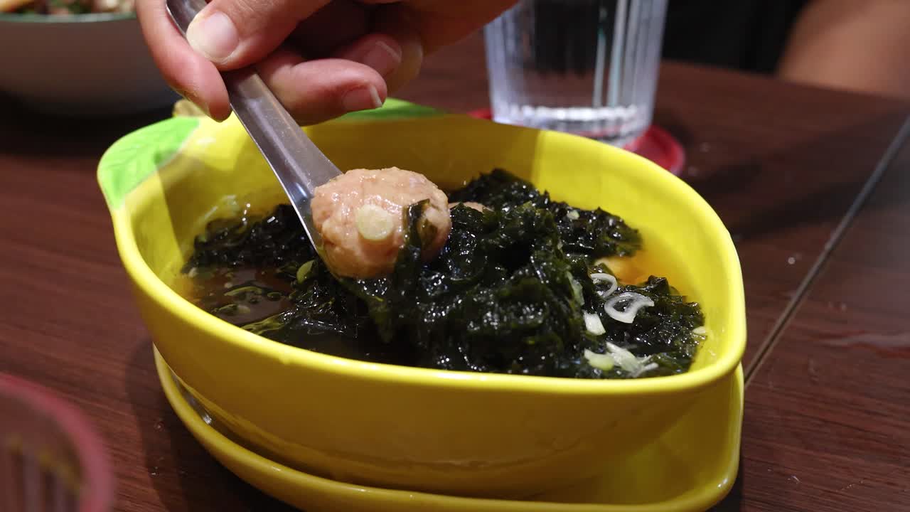 A hand uses a spoon to lift a pork ball and seaweed from a yellow bowl of Thai soup on a wooden table under warm indoor lighting
