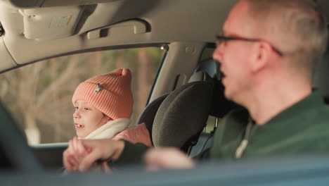 a man is seated in the driver's seat of a car, wearing a green jacket and glasses, smiling and joyfully dancing to music while singing. beside him, a young girl, dressed warmly in a pink jacket