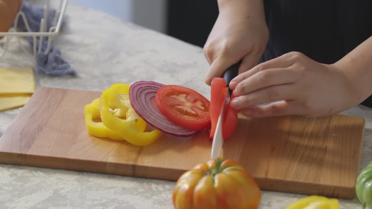 Slicing fresh tomato beside onion and bell pepper on a wooden cutting board, captured in a bright kitchen close-up.