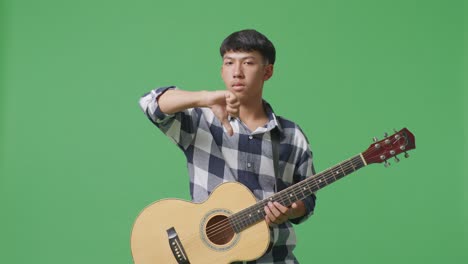 asian teenager with guitar showing thumbs down gesture while standing on green screen background