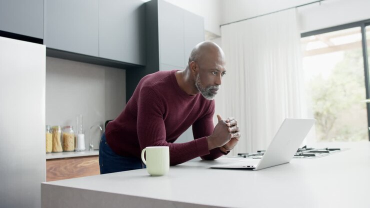 Happy mature biracial man with cup of coffee using laptop in kitchen, slow motion