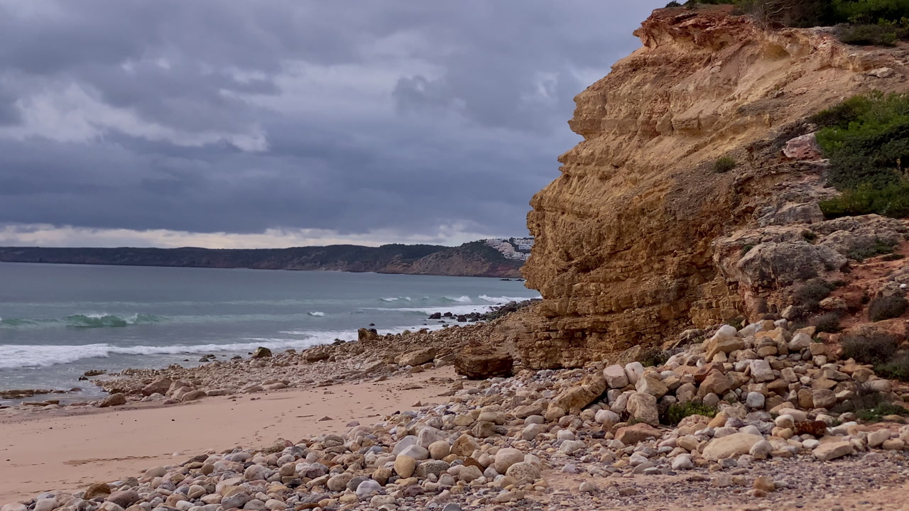 Atlantic waves roll onto a rugged pebble beach beneath sandstone cliffs at Zavial Beach, where clouds and raw textures shape a wild coastal atmosphere