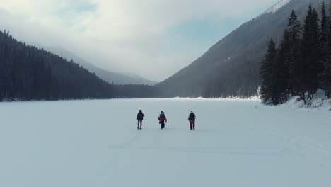 Toma-Aérea-De-Un-Grupo-De-Personas-Caminando-Sobre-Un-Lago-Congelado-Rodeado-De-Pinos-Y-Montañas-Durante-El-Invierno,-Columbia-Británica,-Canadá