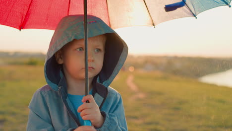 upset kid with umbrella at rainfall on riverbank thoughtful little boy in bad mood stands alone in