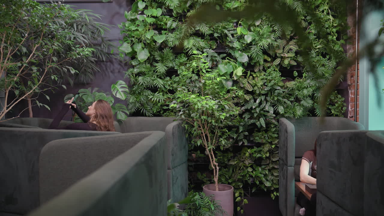 Pretty woman holds phone to photograph greenery while seated in cushioned lounge booth, surrounded by vibrant plants and brick wall, with another person seated across room partially visible