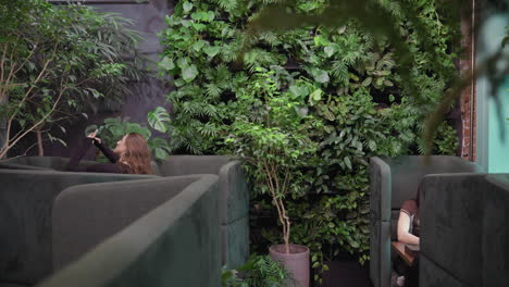 Pretty woman holds phone to photograph greenery while seated in cushioned lounge booth, surrounded by vibrant plants and brick wall, with another person seated across room partially visible