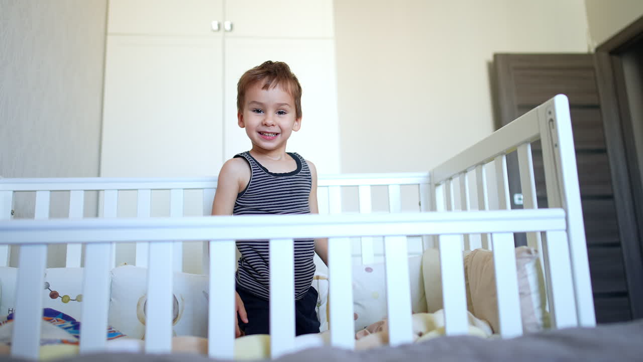 Happy energetic Caucasian toddler sits and hides in his crib. Active baby boy climbs out of bed approaching camera.