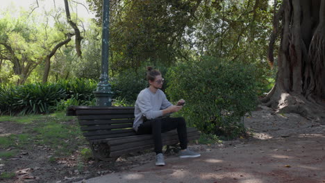 Man-sits-down-on-a-park-bench-and-pulls-out-his-phone,-wide-shot