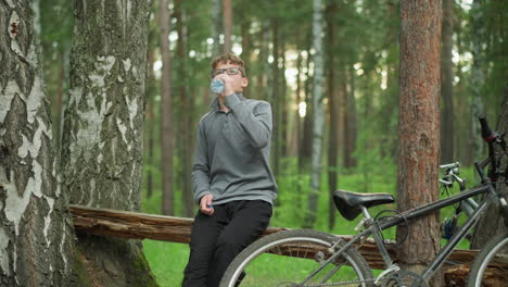 young boy wearing glasses and grey long-sleeve top opens a bottle of water and takes a sip while sitting on a log in the forest, resting beside a parked bicycle surrounded by tall green trees