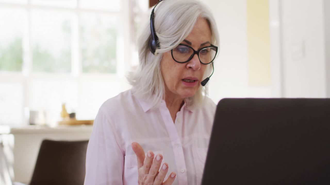 Anziana signora con occhiali che usa un auricolare telefonico mentre fa una videochiamata sul suo laptop mentre lavora