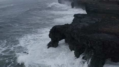 Drone-top-down-of-water-hitting-rocky-cliff-during-cloudy-day-in-Iceland