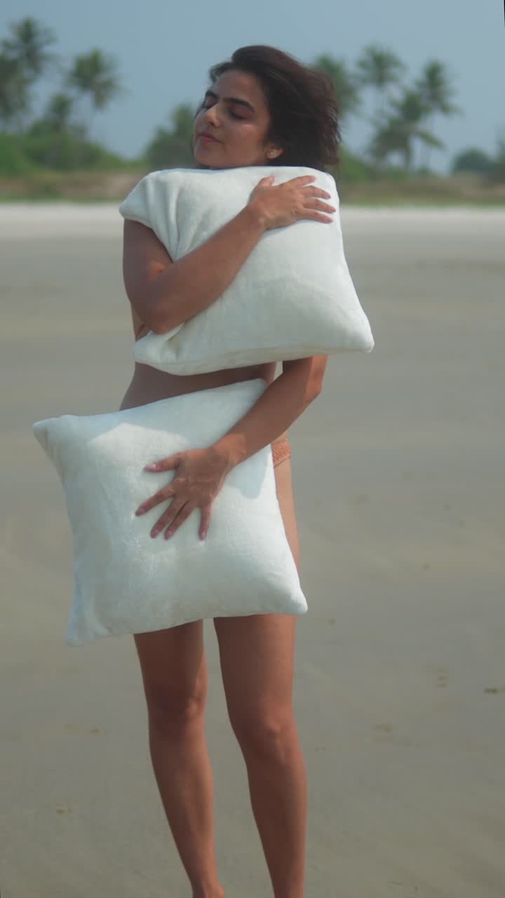 Vertical shot of a woman on the beach holding two white pillows, eyes closed, appearing calm and serene with waves in the background