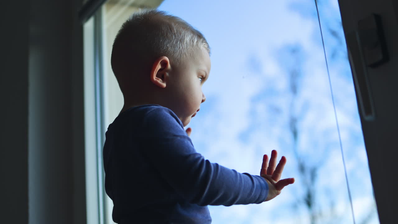 A 1 year old boy is standing on a windowsill and looking out the window. The child looks out the window at the street