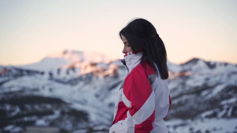 una mujer hermosa con ropa de invierno observa la puesta de sol sobre un grupo de montañas nevadas al atardecer