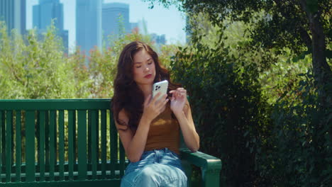 Asian-girl-sitting-on-park-bench-holding-phone.-Woman-dial-phone-number-outdoors