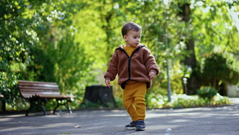 caucasian toddler wearing brown jacket catching soap bubbles. baby boy having fun on the walk in the park.