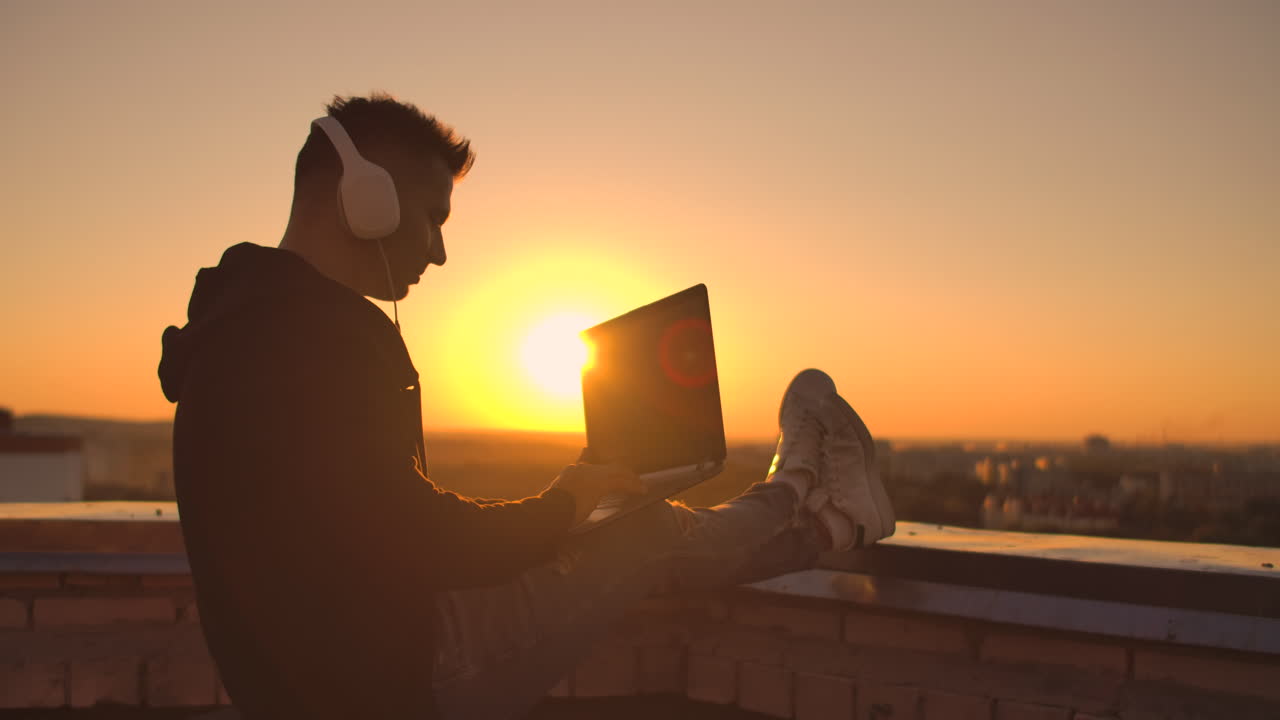 A male freelance programmer sits on a skyscraper roof with a laptop and beer typing code on a keyboard during sunset. Remote work