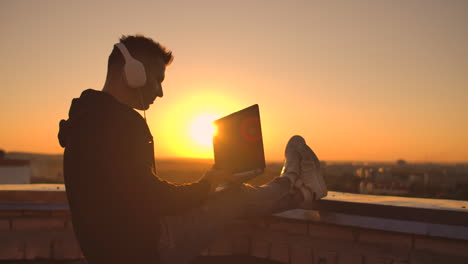 a male freelance programmer sits on a skyscraper roof with a laptop and beer typing code on a keyboard during sunset. remote work