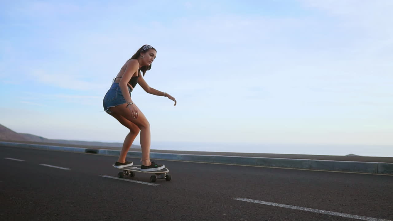 Amid the enchanting sunset hues, a woman skateboards on a road at sunset, captured in slow motion. Mountains and a scenic sky enhance the backdrop, and she's in shorts