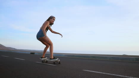 Amid-the-enchanting-sunset-hues,-a-woman-skateboards-on-a-road-at-sunset,-captured-in-slow-motion.-Mountains-and-a-scenic-sky-enhance-the-backdrop,-and-she's-in-shorts