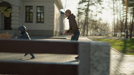 father and son chasing a balloon along a paved path near a building, the child, dressed in a shiny black jacket, reaches out to catch the balloon while the father approaches with open arms