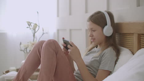 preteen girl is using app for communicating on smartphone talking and listening by headphones resting in her bedroom