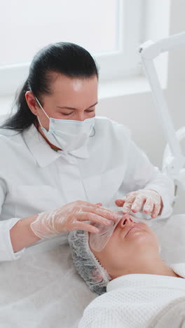 beautician applying regeneration moisturizing sheet mask on young adult woman face skin and eyes