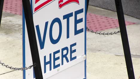 Vote-here-sign-with-American-flag-blowing-in-the-wind,-close-up