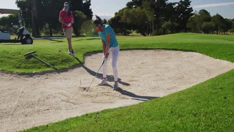 Two-caucasian-women-playing-golf-wearing-face-masks-one-taking-shot-from-bunker