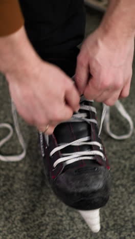 primer plano de las manos atando los cordones de un patín de hielo negro con acentos rojos, la persona lleva pantalones negros y una camiseta de manga marrón