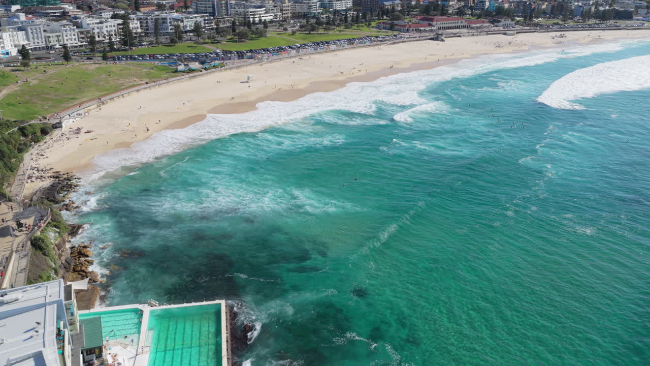Una ripresa aerea dinamica inclinata verso l'alto, sopra l'acqua turchese e la sabbia della pittoresca Bondi Beach, a Sydney, in Australia, con surfisti che cavalcano le onde bianche e nuotatori e turisti che si godono la giornata di sole