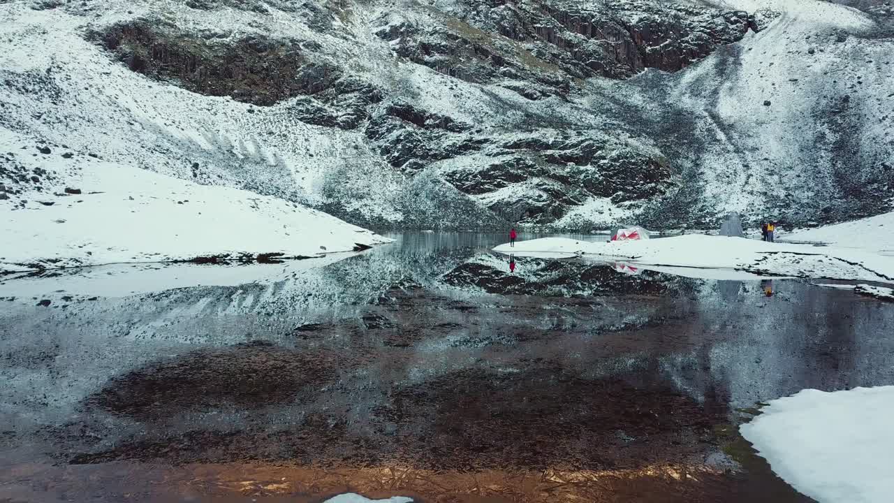 Aerial, drone shot over a reflecting glacier lake, people on the shore, in Andes mountains, on a snowy day, in Cusco region, Peru