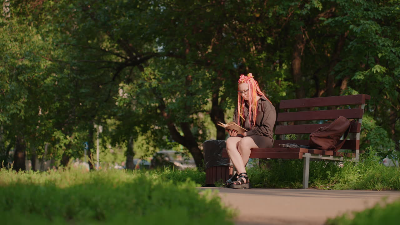 Женская фигура тихо сидит среди деревьев вечером, одинокая женщина отдыхает на скамейке возле зеленой листвы на закате, уединенная женщина задумчиво сидит на скамейке среди деревьев в сумерках