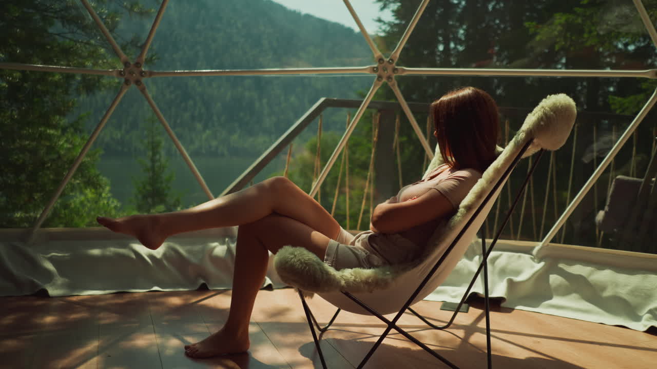 Depressed woman sits in armchair with fur near panoramic window in tourist area overlooking dense forest and lake. Relaxation in solitude and peace with nature