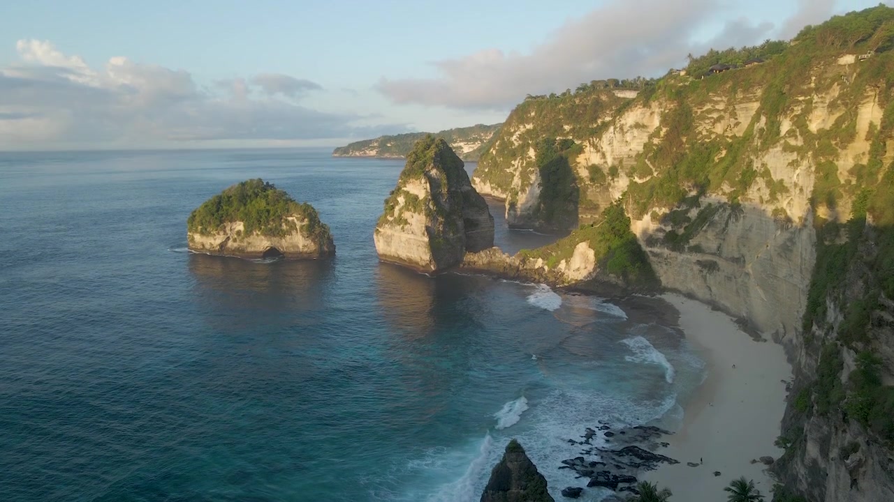 Eine atemberaubende filmische Drohnenaufnahme vom Diamond Beach in Nusa Penida, Bali, mit Kalksteinfelsen, türkisfarbenem Wasser und ikonischen Felsformationen bei Sonnenaufgang.