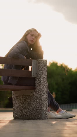 dama sentada en el borde de un banco del parque durante la hora dorada, descansando la cabeza de una manera contemplativa, mientras la luz del sol brilla brillantemente, con una figura borrosa caminando en el fondo