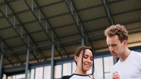 joven deportivo revisando el reloj inteligente de su novia, mientras tiene una conversación divertida con ella y bebe agua en el estadio