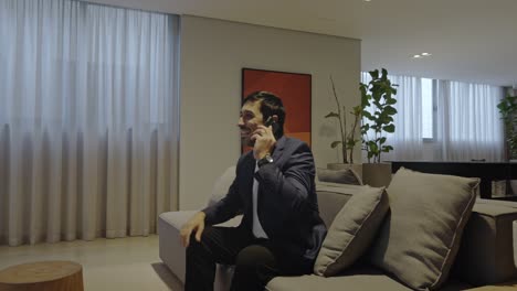 executive man sitting on a comfortable sofa smiling and talking on the phone inside a modern living room.