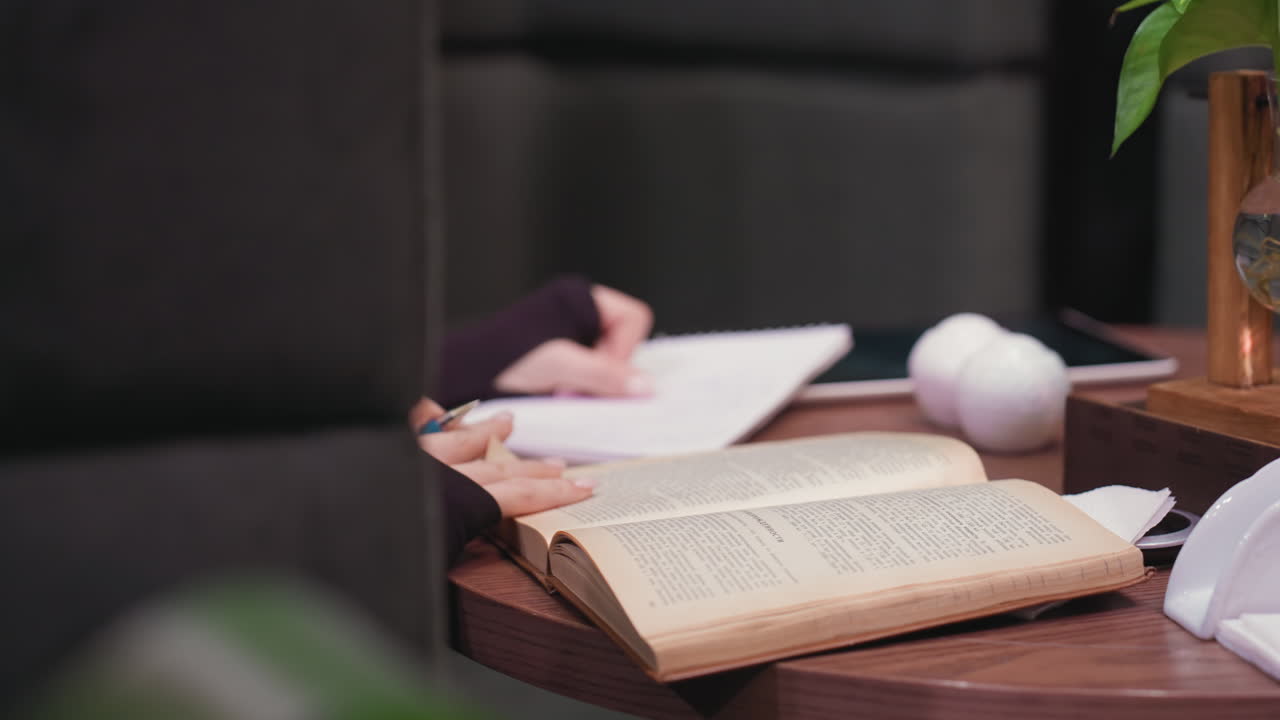 Close up of woman hand holding pen while turning page of worn brown book on wooden table, with blurred notebook, white decoration, and green plant in background creating focused learning ambiance