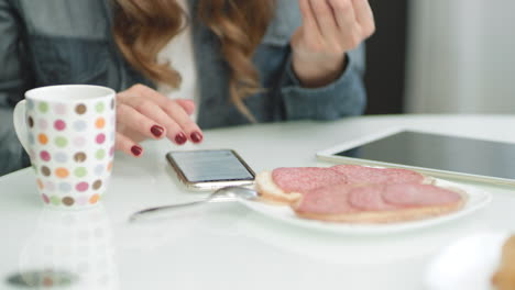 Closeup-female-hands-scrolling-mobile-phone-while-eating-sandwich